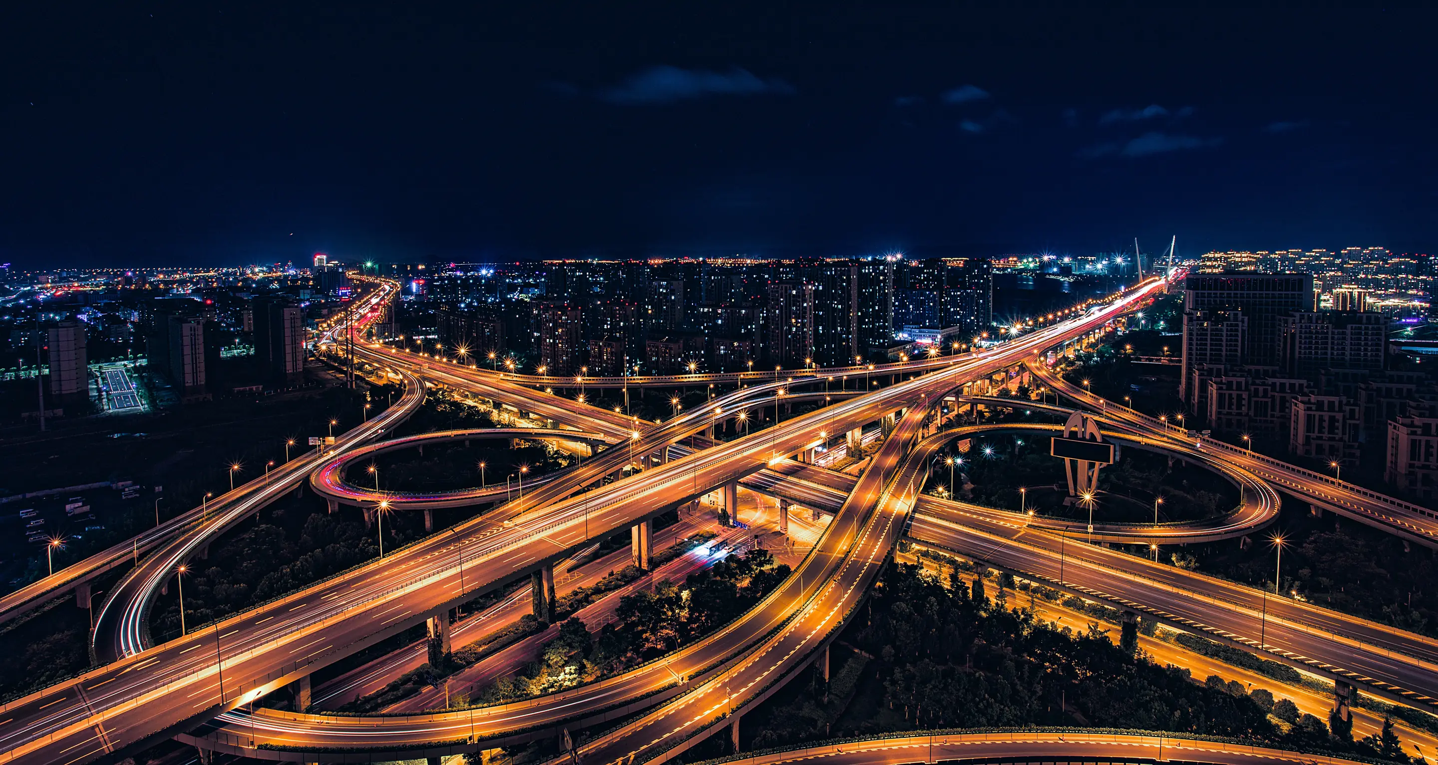 Aerial night view of a multi-level motorway interchange, its looping ramps traced in amber light against a dark city